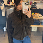 Person taking a mirror selfie wearing a brown jacket and blue jeans in a store.