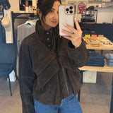 Person taking a mirror selfie wearing a brown jacket and blue jeans in a store.