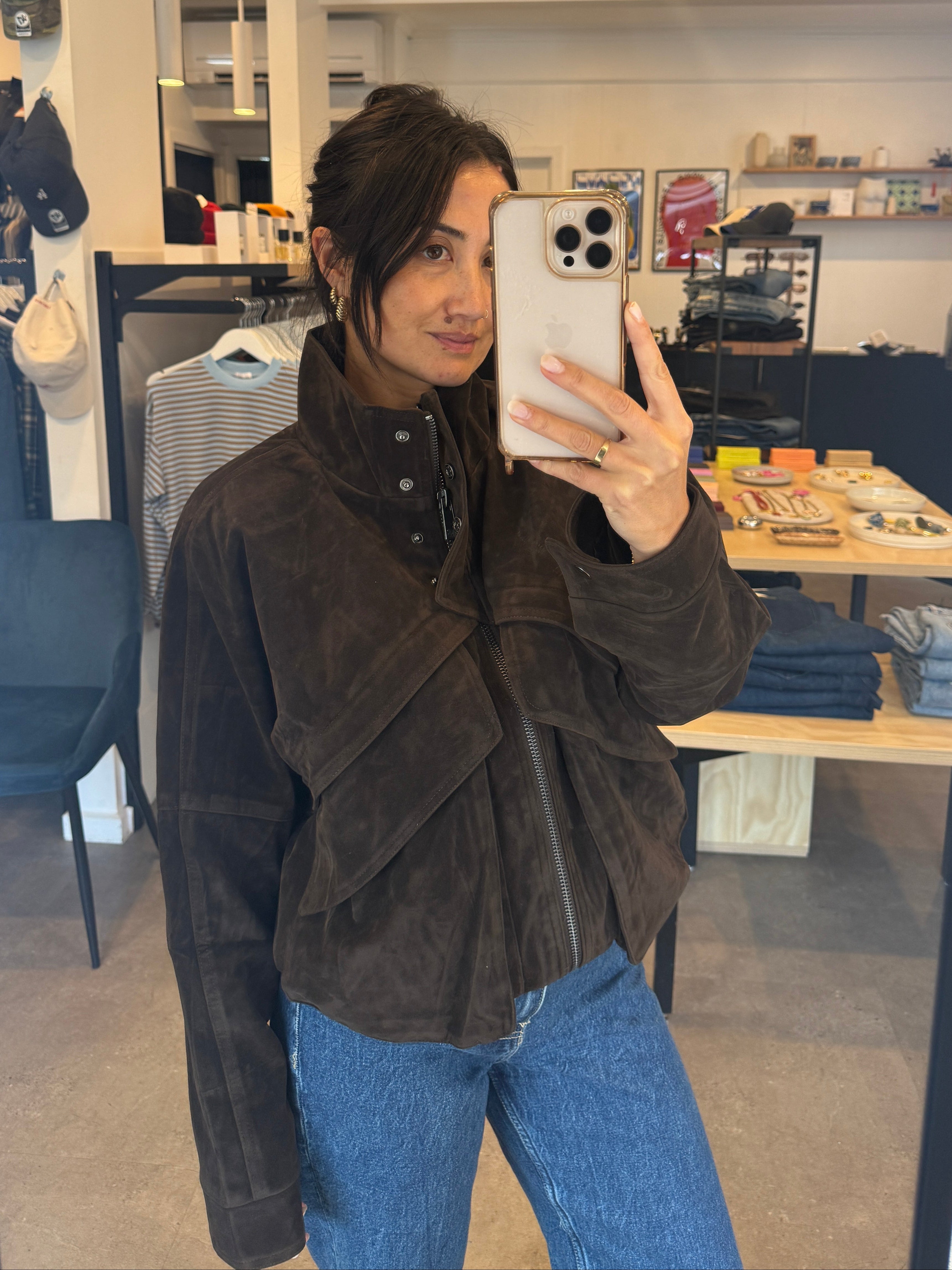 Person taking a mirror selfie wearing a brown jacket and blue jeans in a store.