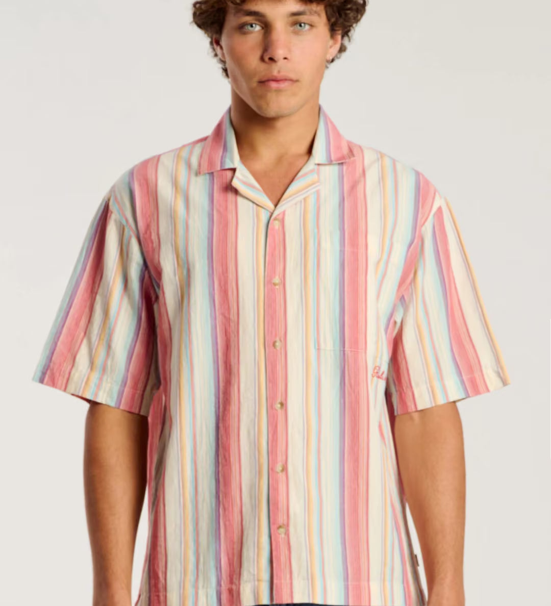 A man wearing a short-sleeve button-up shirt with vertical pastel stripes in pink, orange, yellow, blue, and white, paired with blue jeans, against a light gray studio background.