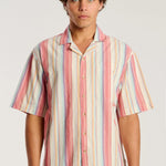 A man wearing a short-sleeve button-up shirt with vertical pastel stripes in pink, orange, yellow, blue, and white, paired with blue jeans, against a light gray studio background.