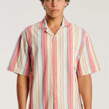A man wearing a short-sleeve button-up shirt with vertical pastel stripes in pink, orange, yellow, blue, and white, paired with blue jeans, against a light gray studio background.