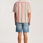 A man viewed from behind wearing a short-sleeve button-up shirt with vertical stripes in pink, orange, yellow, and blue on a cream base, paired with denim shorts and tan sandals, against a plain white studio background.