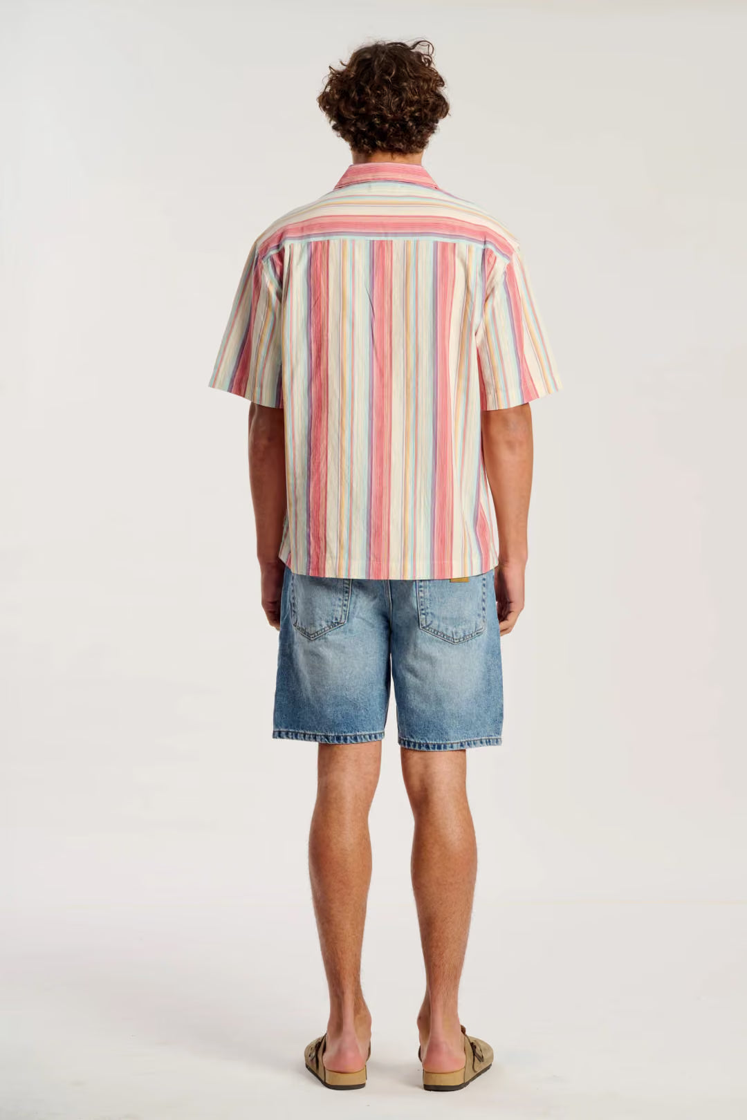 A man viewed from behind wearing a short-sleeve button-up shirt with vertical stripes in pink, orange, yellow, and blue on a cream base, paired with denim shorts and tan sandals, against a plain white studio background.