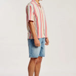 A man wearing a short-sleeve button-up shirt with vertical pink, red, and cream stripes, paired with light blue denim shorts and tan suede mule loafers, standing in profile against a plain light gray studio background.
