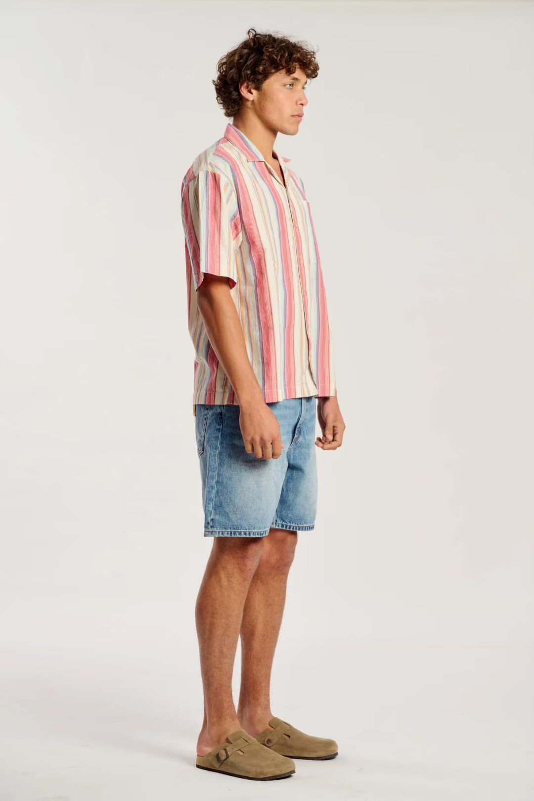 A man wearing a short-sleeve button-up shirt with vertical pink, red, and cream stripes, paired with light blue denim shorts and tan suede mule loafers, standing in profile against a plain light gray studio background.