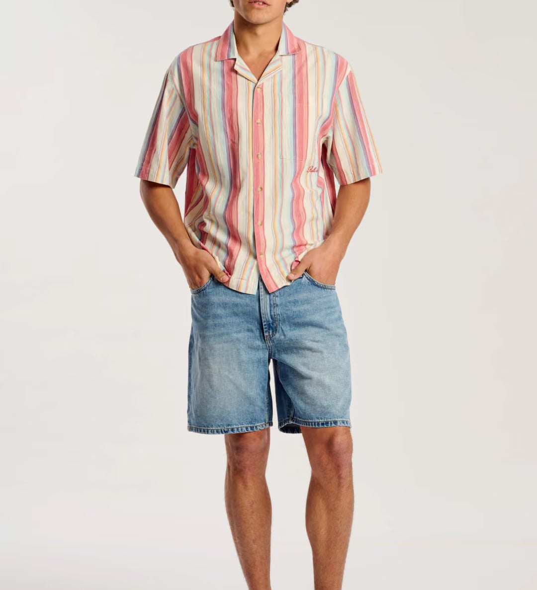 A man modeling a short-sleeve button-up shirt with vertical pink, orange, yellow, and white stripes, paired with blue denim shorts and tan suede mules, against a plain white studio background.