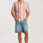 A man modeling a short-sleeve button-up shirt with vertical pink, orange, yellow, and white stripes, paired with blue denim shorts and tan suede mules, against a plain white studio background.
