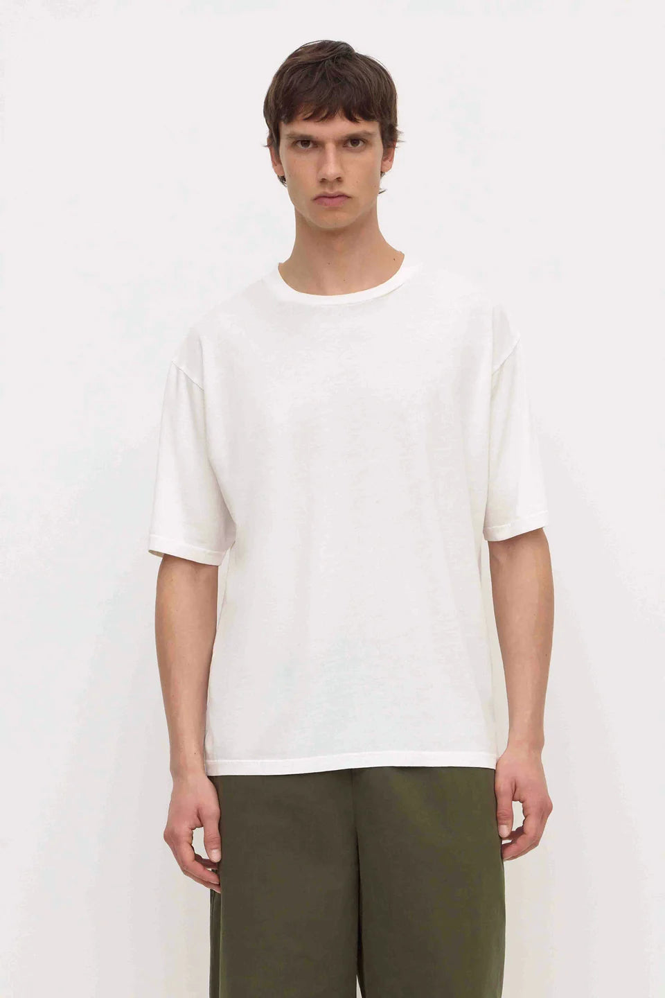 A man wearing a plain white short-sleeve crewneck t-shirt and olive green pants, standing against a light off-white background.