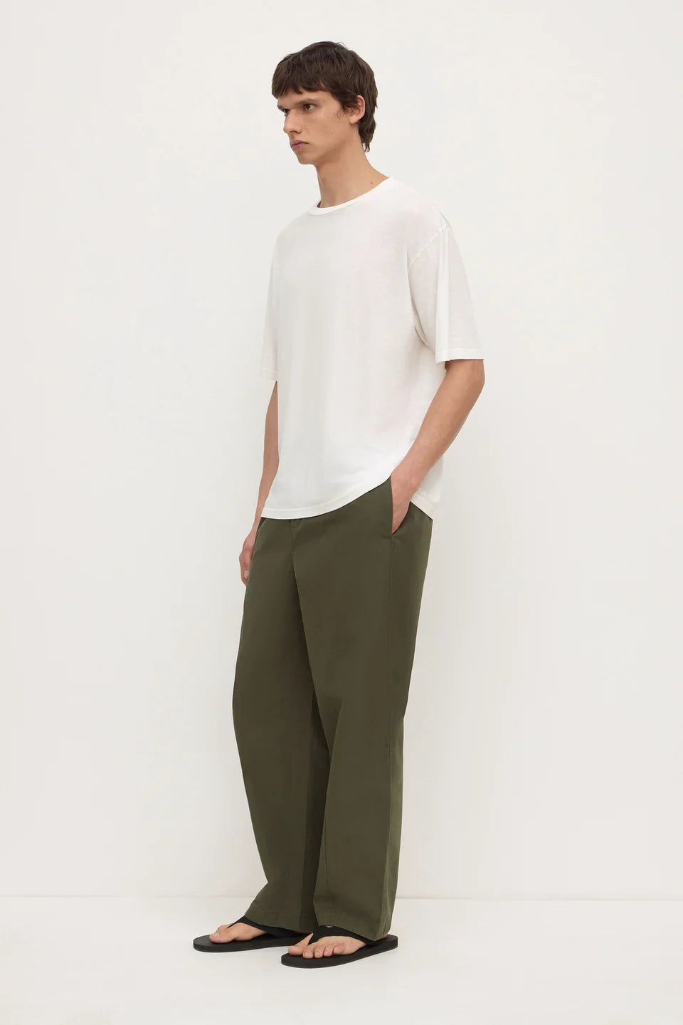 A man wearing a white short-sleeve crewneck t-shirt, olive green wide-leg trousers, and black flip-flops, standing against a plain off-white studio background.