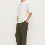 A man wearing a white short-sleeve crewneck t-shirt, olive green wide-leg trousers, and black flip-flops, standing against a plain off-white studio background.