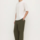 A man wearing a white short-sleeve crewneck t-shirt, olive green wide-leg trousers, and black flip-flops, standing against a plain off-white studio background.