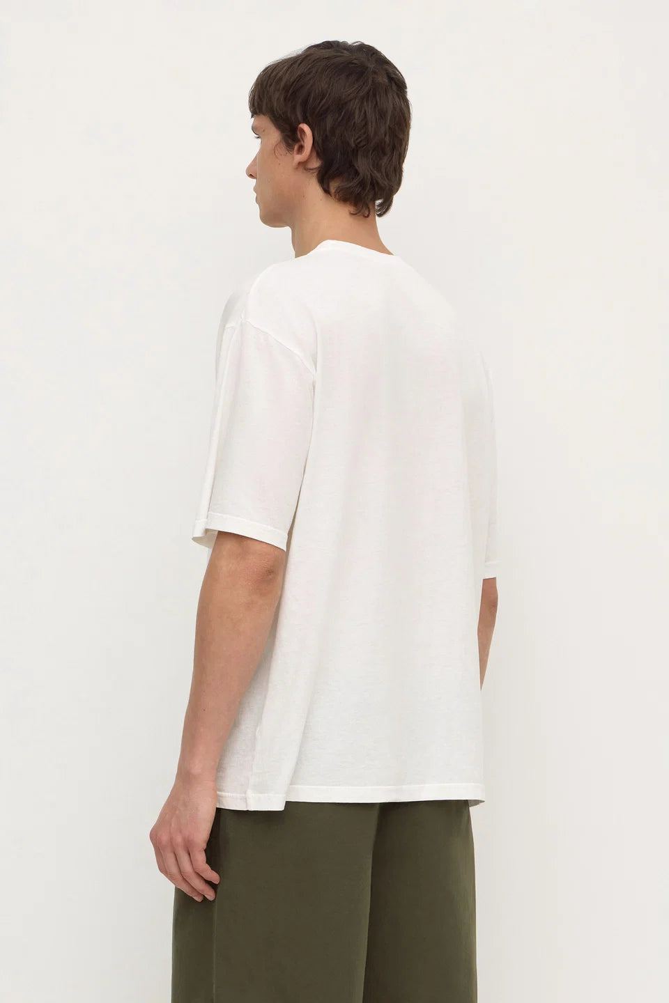 A man shown from the back wearing an oversized white short-sleeve t-shirt and olive green pants, standing against a plain off-white studio background.