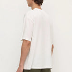 A man shown from the back wearing an oversized white short-sleeve t-shirt and olive green pants, standing against a plain off-white studio background.