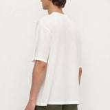 A man shown from the back wearing an oversized white short-sleeve t-shirt and olive green pants, standing against a plain off-white studio background.