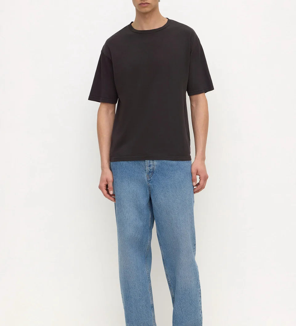A man modeling a black short-sleeve crewneck t-shirt, paired with light blue relaxed-fit jeans and black sandals, standing against a plain white studio background.