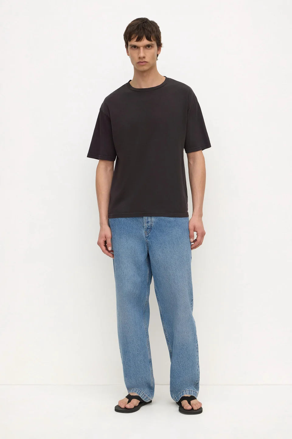 A man modeling a black short-sleeve crewneck t-shirt, paired with light blue relaxed-fit jeans and black sandals, standing against a plain white studio background.