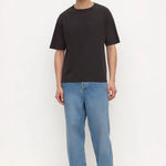 A man modeling a black short-sleeve crewneck t-shirt, paired with light blue relaxed-fit jeans and black sandals, standing against a plain white studio background.