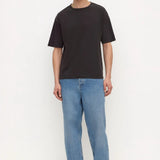 A man modeling a black short-sleeve crewneck t-shirt, paired with light blue relaxed-fit jeans and black sandals, standing against a plain white studio background.