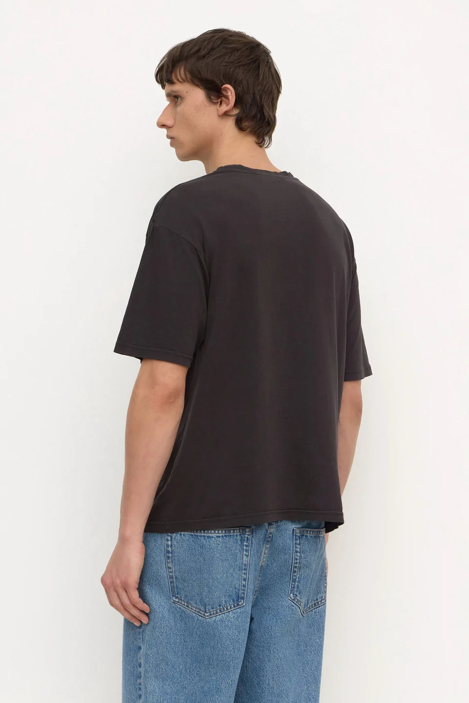 A man wearing a dark charcoal gray oversized t-shirt and blue jeans, shown from a side-back angle against a plain white studio background.