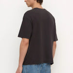 A man wearing a dark charcoal gray oversized t-shirt and blue jeans, shown from a side-back angle against a plain white studio background.