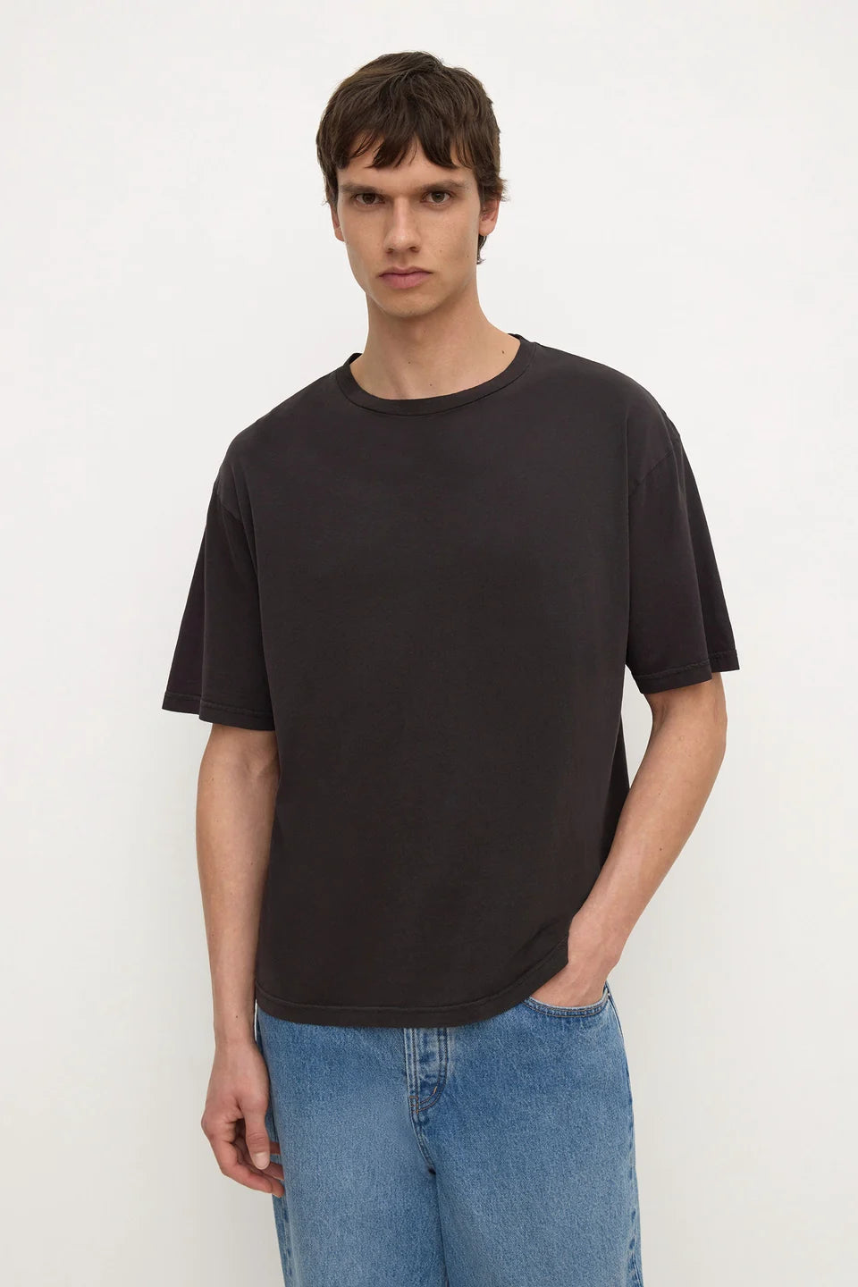 A man wearing a dark charcoal gray crewneck t-shirt and light blue jeans, standing against a plain off-white background.