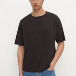 A man wearing a dark charcoal gray crewneck t-shirt and light blue jeans, standing against a plain off-white background.