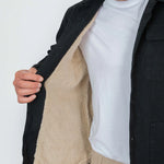 Person wearing a black coat with a beige fleece lining, white shirt, and beige pants on a white background.