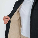 Person wearing a black coat with a beige fleece lining, white shirt, and beige pants on a white background.