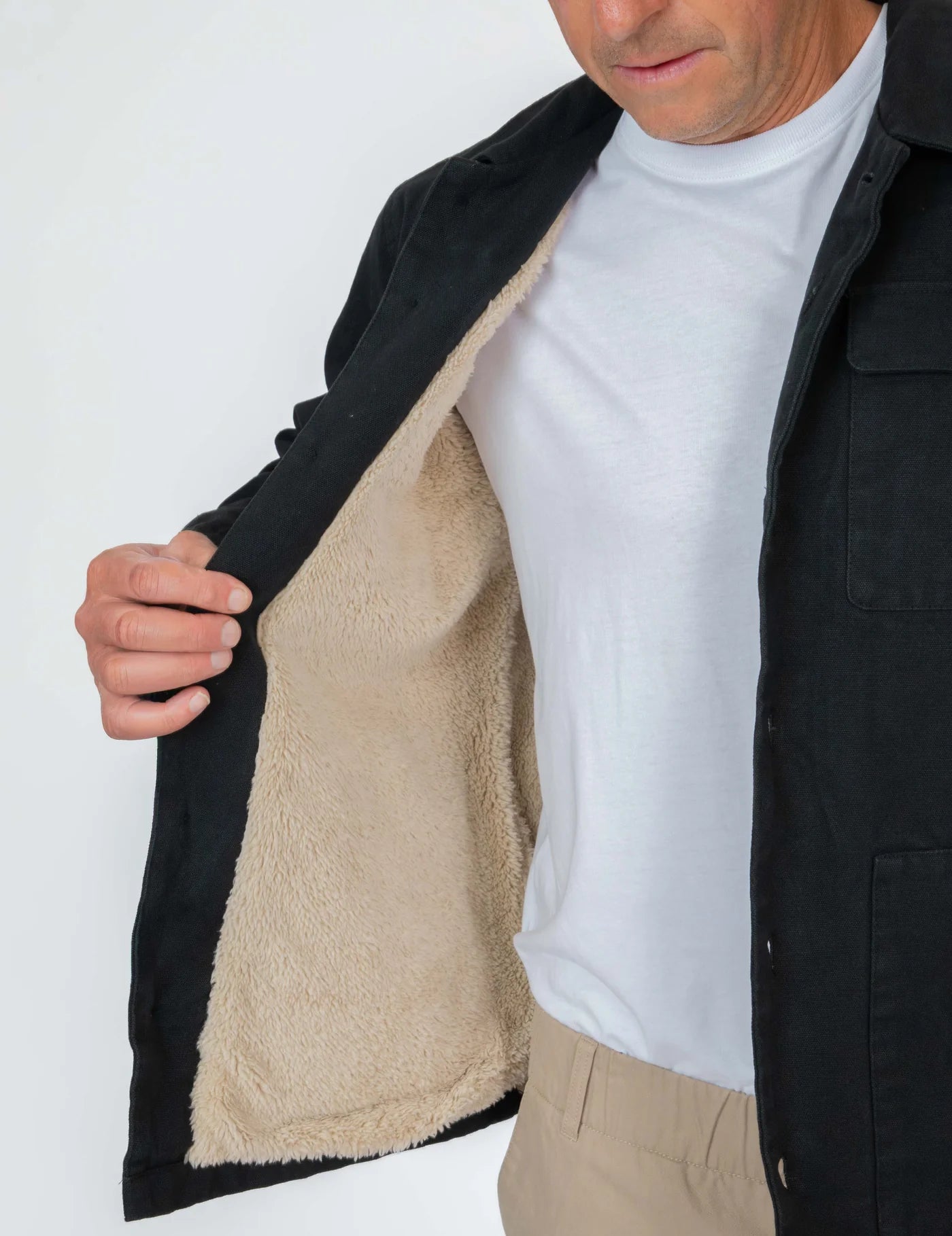 Person wearing a black coat with a beige fleece lining, white shirt, and beige pants on a white background.
