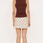 A woman shown from the back wearing a sleeveless brown ribbed knit top and a cream mini skirt with brown polka dots, standing against a plain off-white background.
