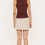 A woman shown from the back wearing a sleeveless brown ribbed knit top and a cream mini skirt with brown polka dots, standing against a plain off-white background.