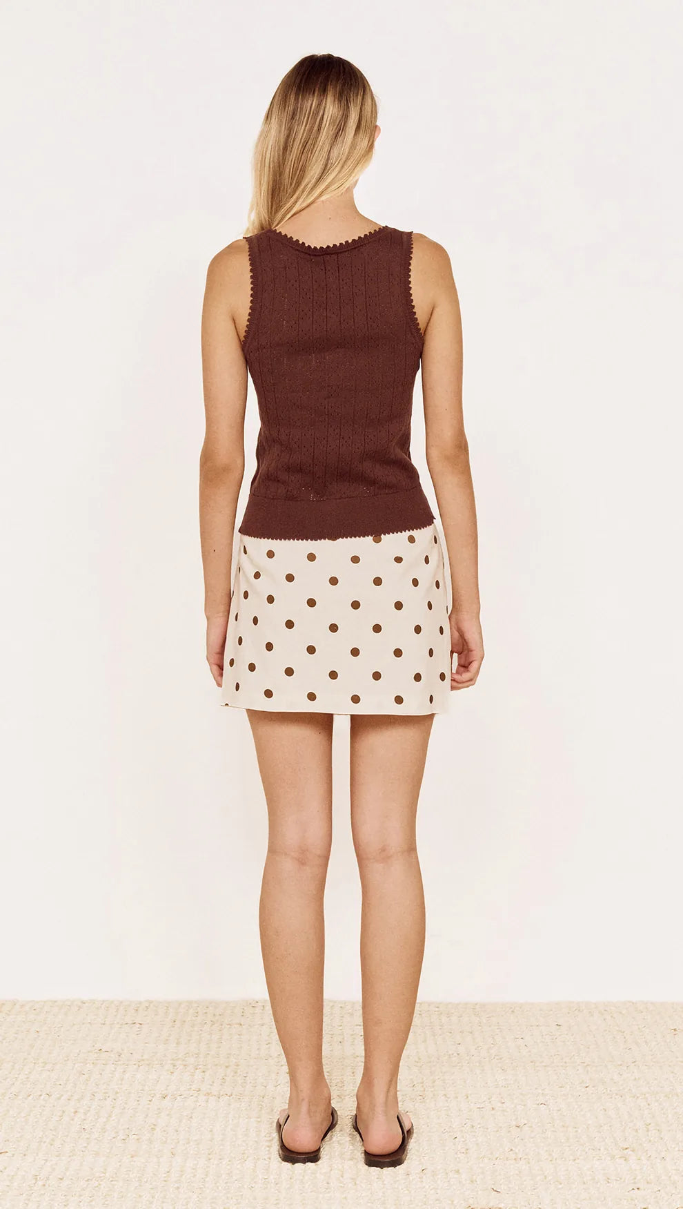 A woman shown from the back wearing a sleeveless brown ribbed knit top and a cream mini skirt with brown polka dots, standing against a plain off-white background.