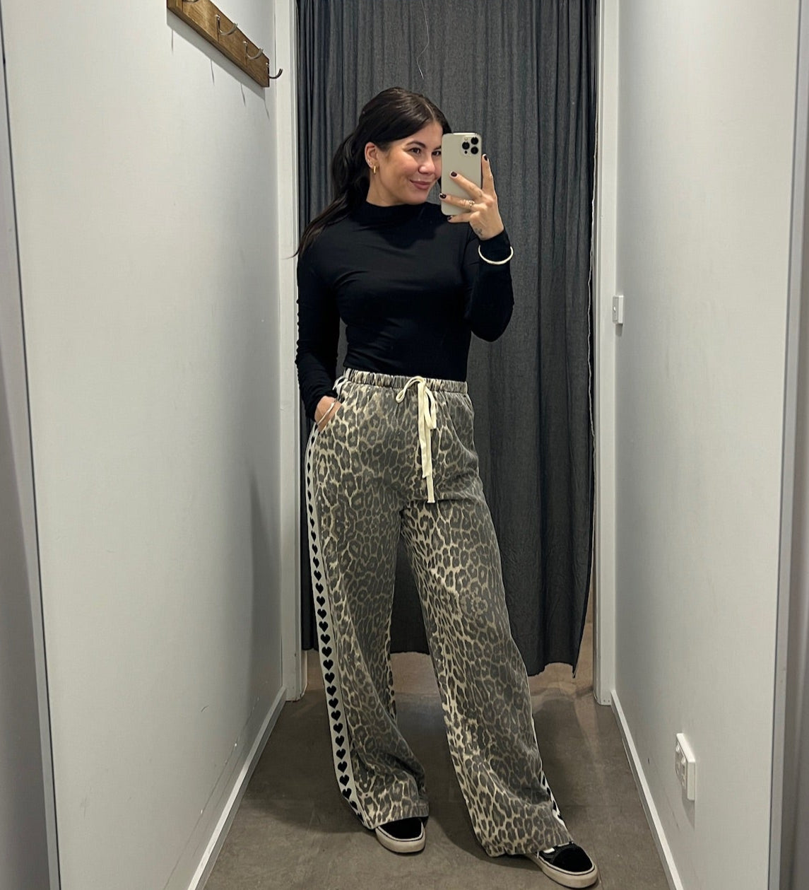 Person taking a mirror selfie wearing a black top and patterned pants in a hallway.