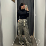 Person taking a mirror selfie wearing a black top and patterned pants in a hallway.
