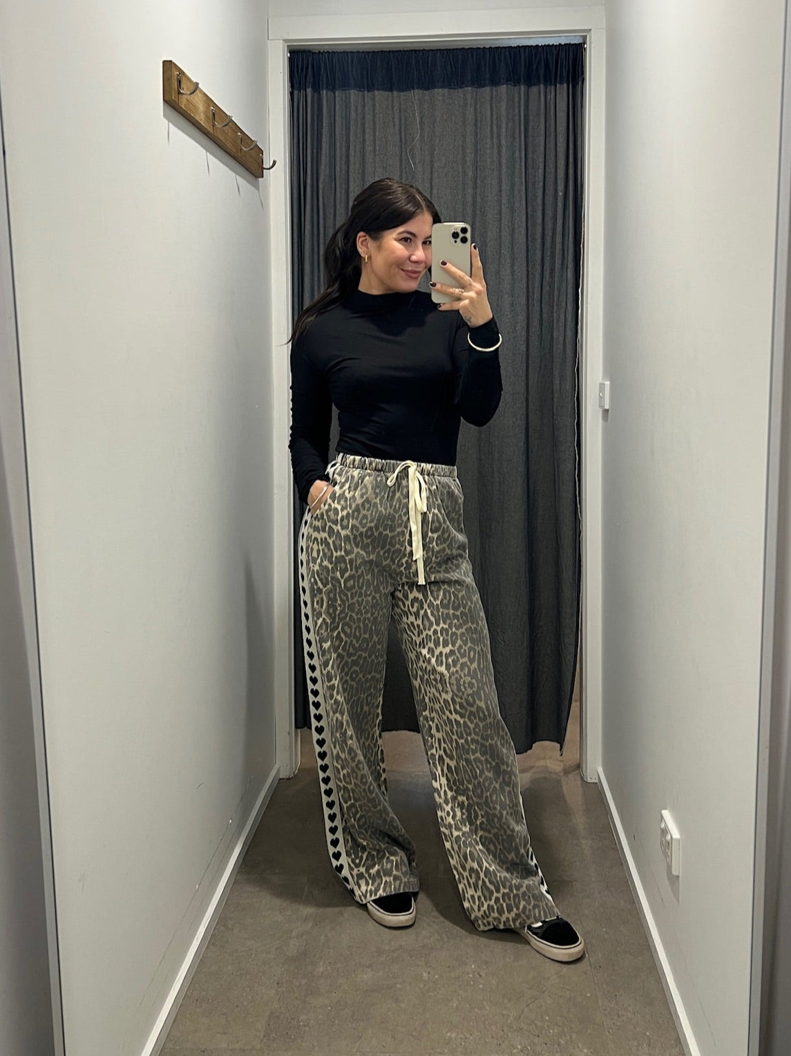 Person taking a mirror selfie wearing a black top and patterned pants in a hallway.