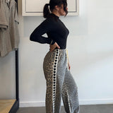 Person wearing a black top and patterned pants standing in a room with a rack of clothes and a whiteboard on the wall.