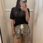 A woman takes a mirror selfie in a fitting room wearing a black short-sleeve t-shirt and leopard print shorts with a cream drawstring.