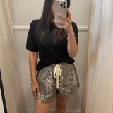 A woman takes a mirror selfie in a fitting room wearing a black short-sleeve t-shirt and leopard print shorts with a cream drawstring.