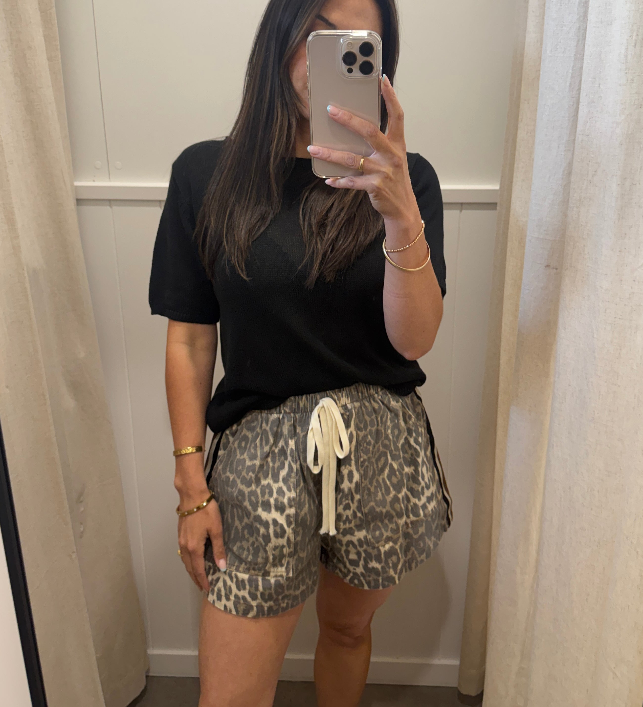 A woman takes a mirror selfie in a fitting room wearing a black short-sleeve t-shirt and leopard print shorts with a cream drawstring.