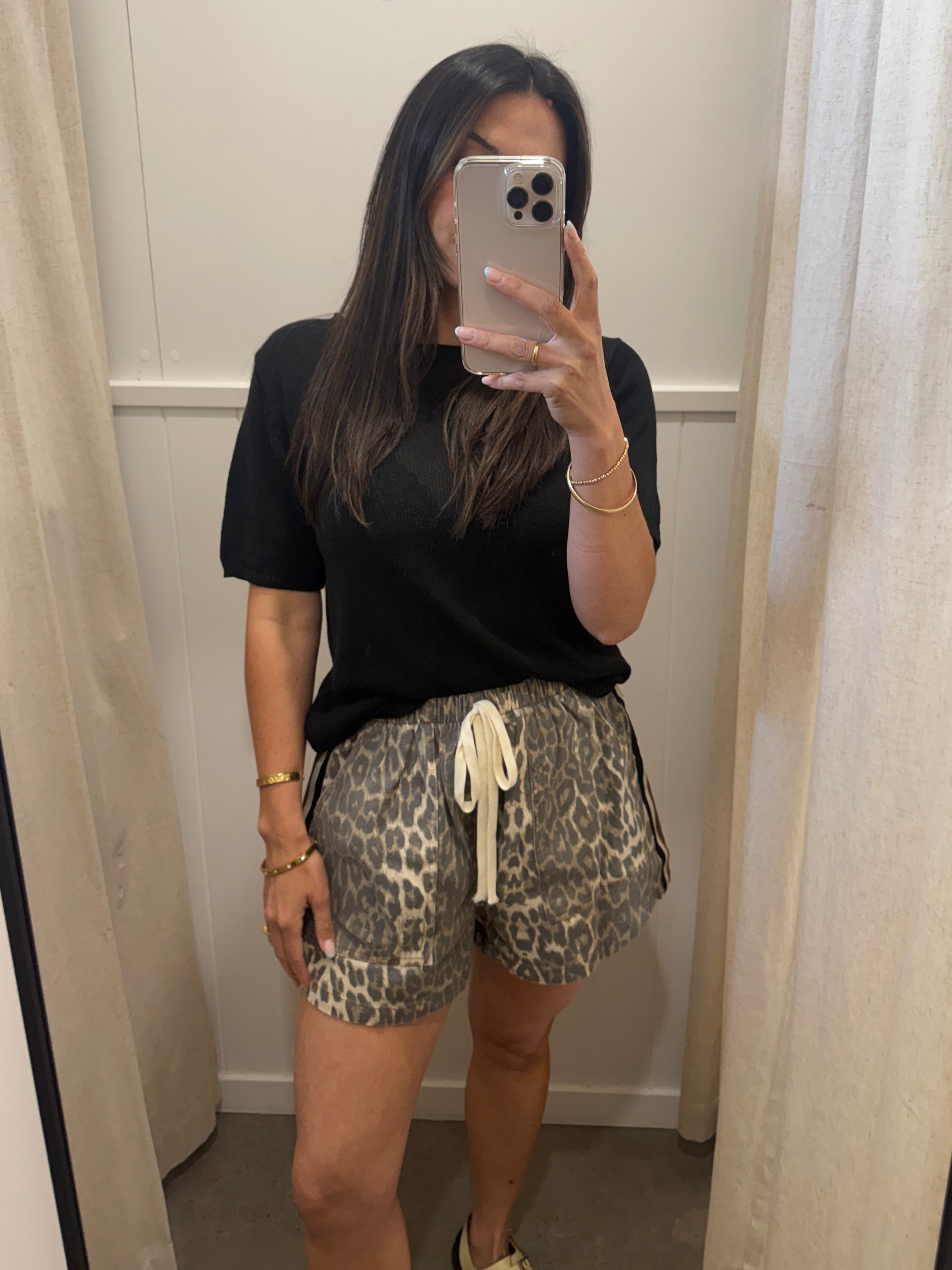 A woman takes a mirror selfie in a fitting room wearing a black short-sleeve t-shirt and leopard print shorts with a cream drawstring.