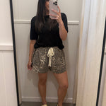 A woman takes a mirror selfie in a fitting room wearing a black short-sleeve t-shirt, leopard-print shorts with a cream drawstring, and beige slide sandals.
