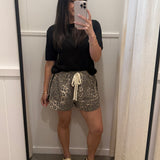 A woman takes a mirror selfie in a fitting room wearing a black short-sleeve t-shirt, leopard-print shorts with a cream drawstring, and beige slide sandals.