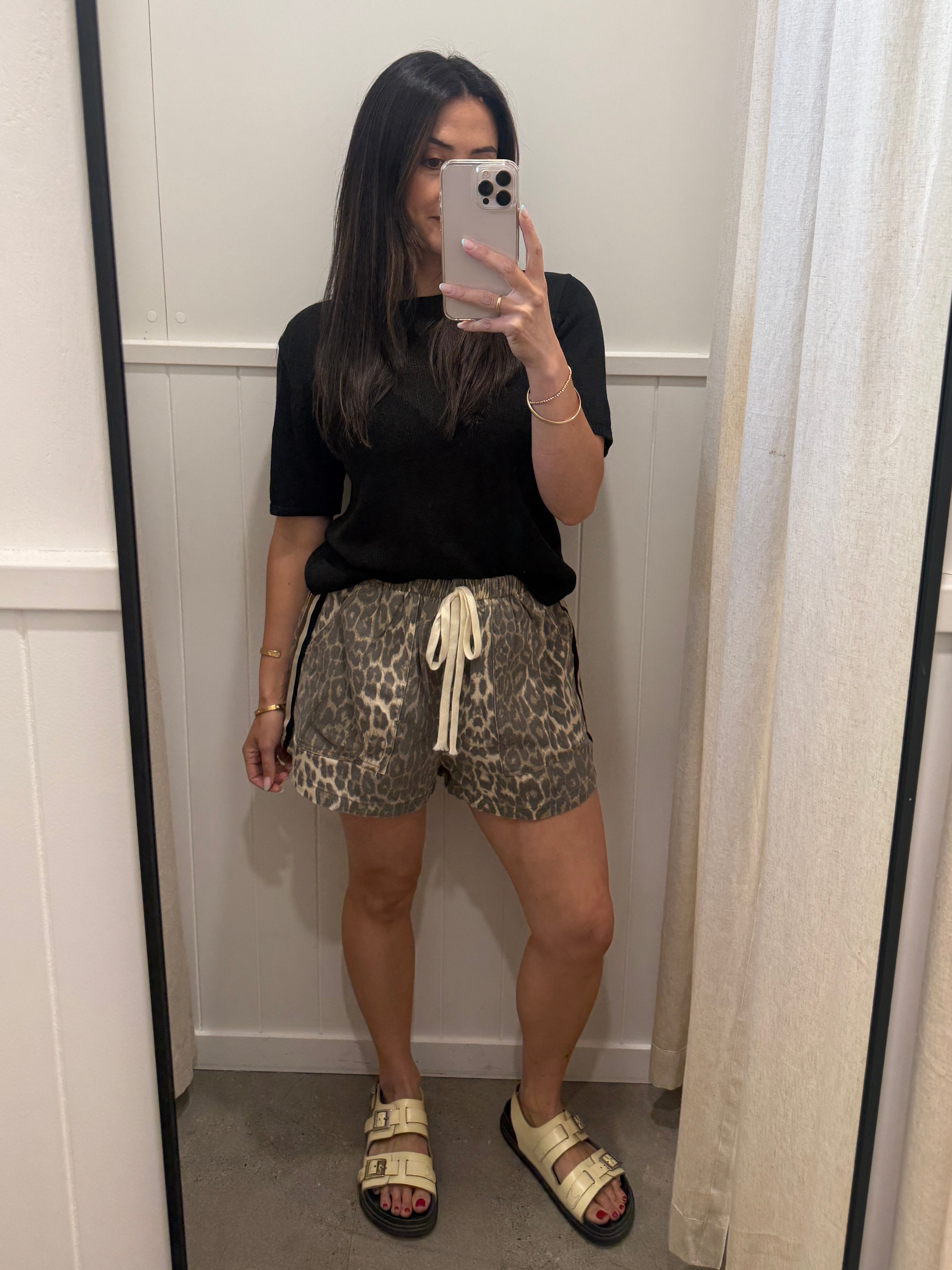 A woman takes a mirror selfie in a fitting room wearing a black short-sleeve t-shirt, leopard-print shorts with a cream drawstring, and beige slide sandals.