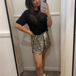 A woman takes a mirror selfie in a fitting room wearing a black short-sleeve t-shirt and leopard-print shorts with a cream drawstring, paired with beige slide sandals.