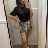 A woman takes a mirror selfie in a fitting room wearing a black short-sleeve t-shirt and leopard-print shorts with a cream drawstring, paired with beige slide sandals.