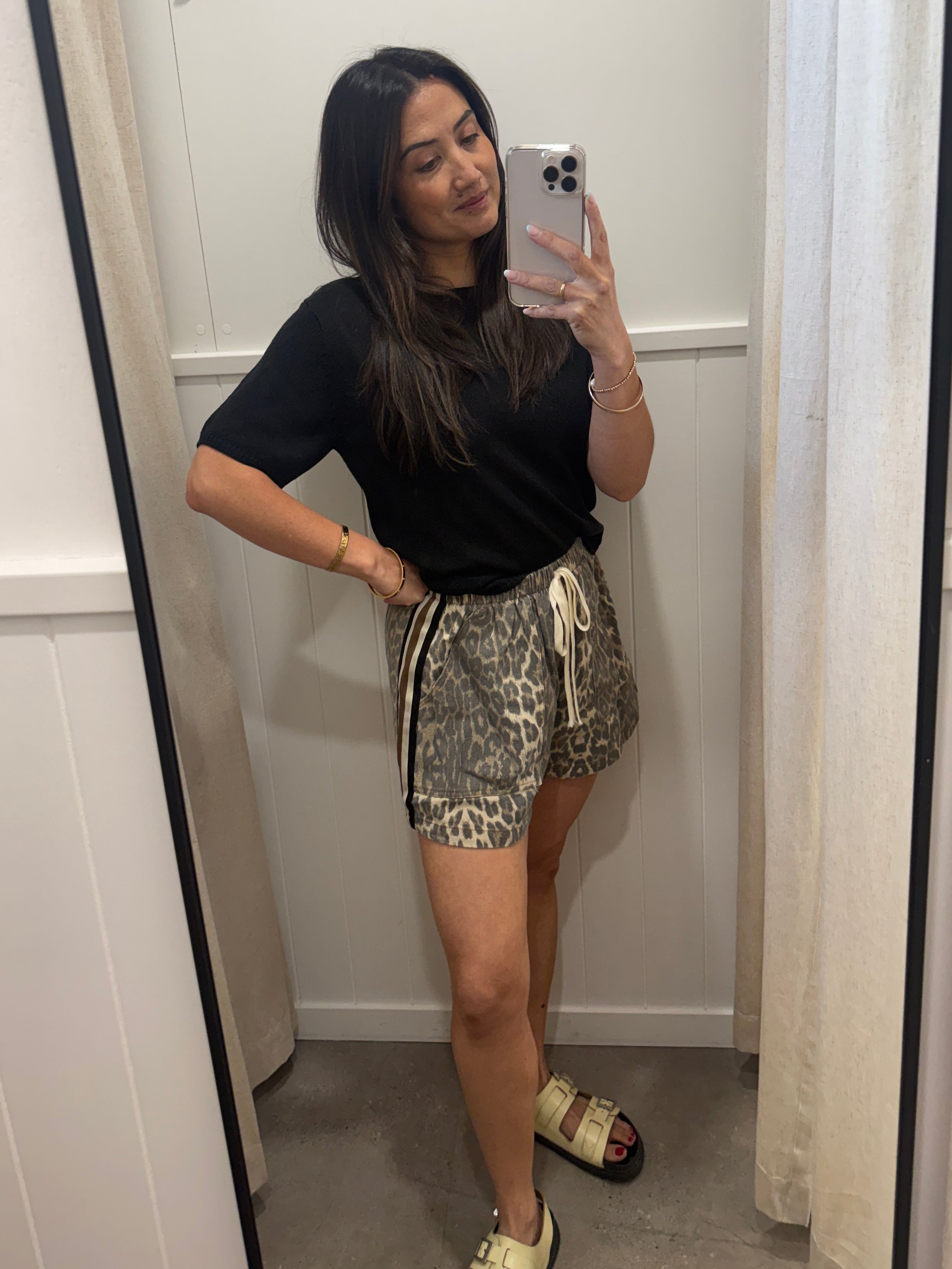A woman takes a mirror selfie in a fitting room wearing a black short-sleeve t-shirt and leopard-print shorts with a cream drawstring, paired with beige slide sandals.