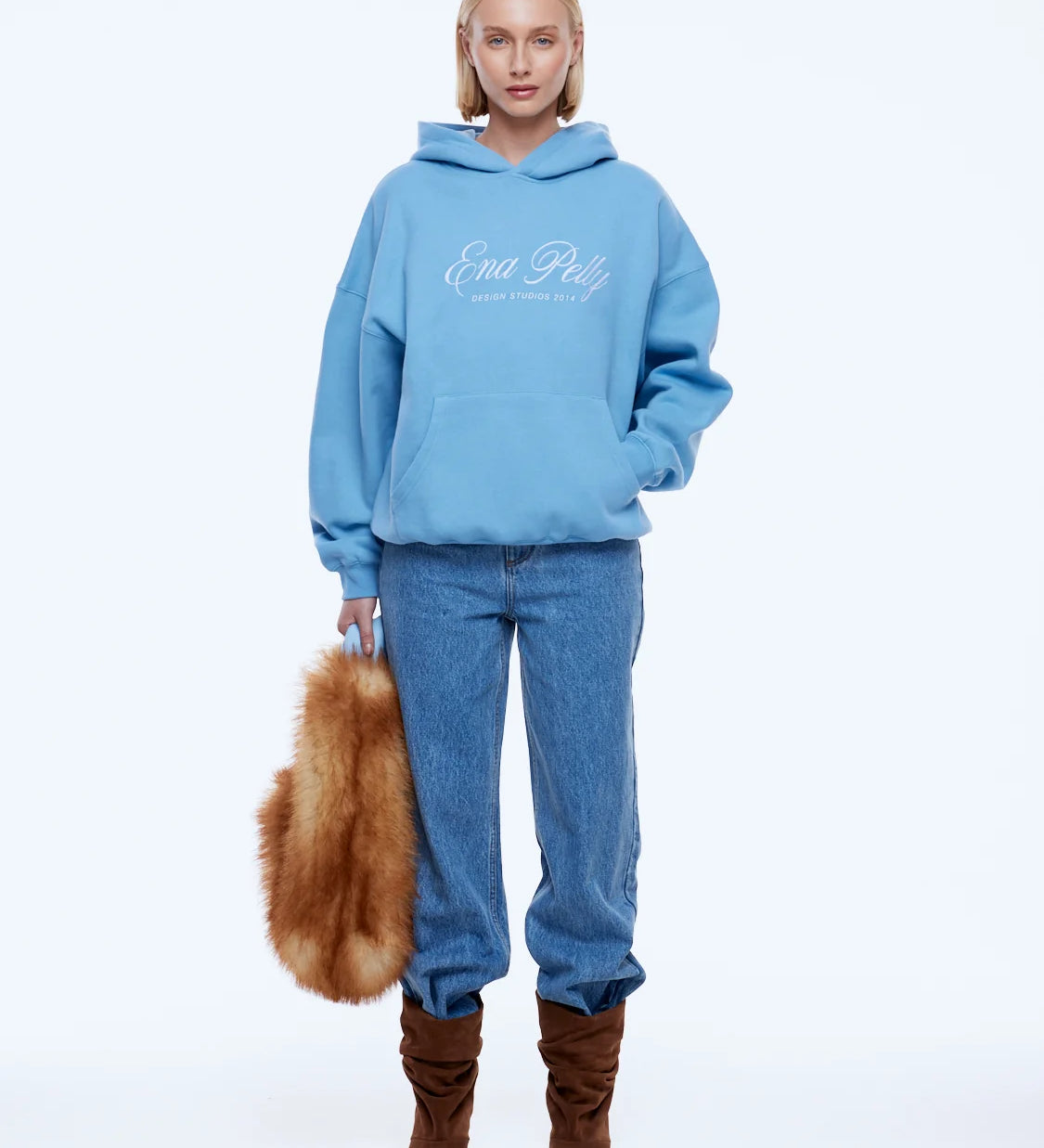 A model stands against a plain white studio background wearing a light blue hoodie with white 'Ena Pelly' script on the chest, blue jeans, and brown boots, while holding a brown fur accessory.