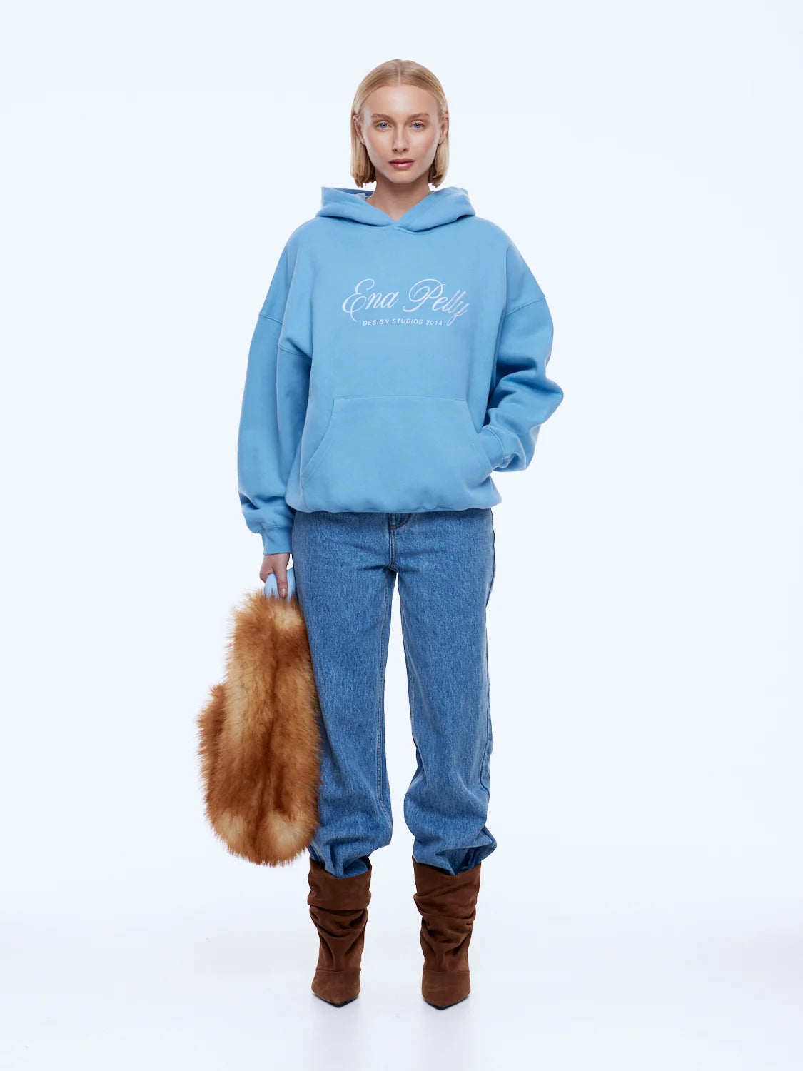 A model stands against a plain white studio background wearing a light blue hoodie with white 'Ena Pelly' script on the chest, blue jeans, and brown boots, while holding a brown fur accessory.