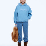 A model stands against a plain white studio background wearing a light blue hoodie with white 'Ena Pelly' script on the chest, blue jeans, and brown boots, while holding a brown fur accessory.